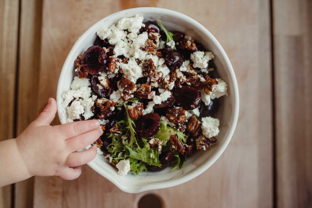 a bowl of cherry goat cheese salad on a wooden cutting board with a baby's hand reaching in