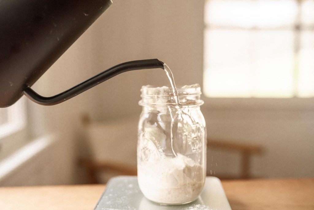 a black kettle pouring water into a glass jar with sourdough starter