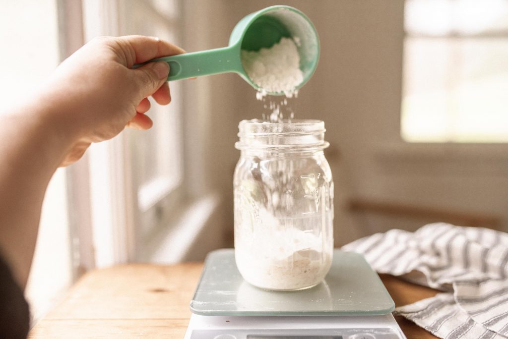 hand holding measuring cup of flour pouring into a glass of sourdough starter