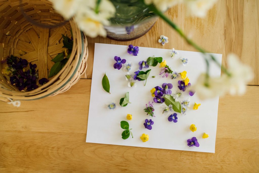 aerial view of a basket with flowers, a paper with violets and buttercups and leaves with a vase of gardenias in the background. 