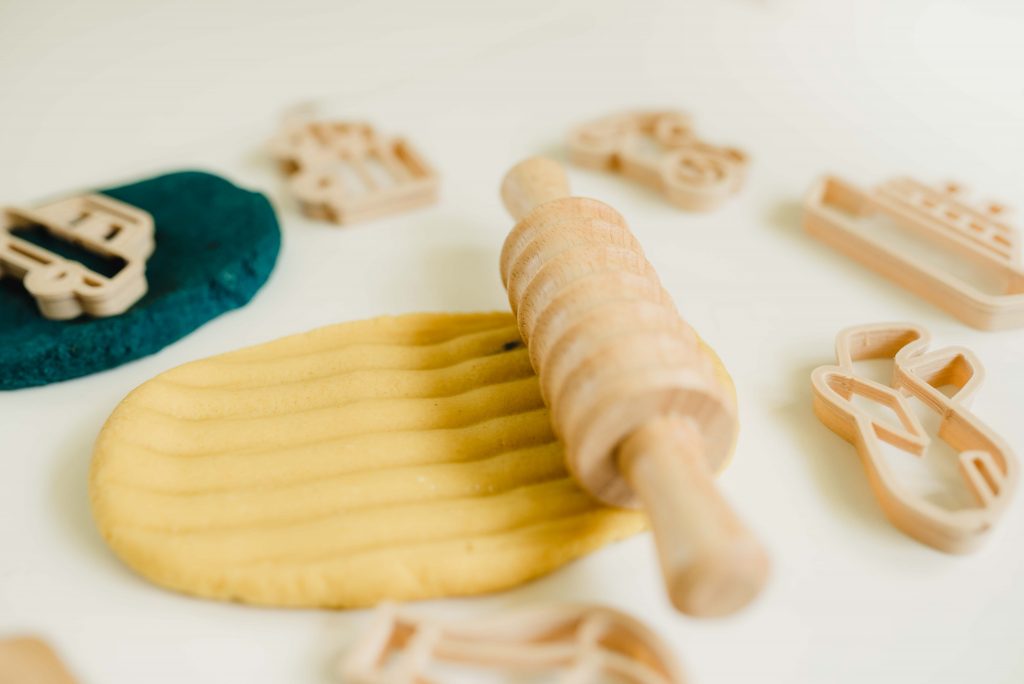 a white table of non-toxic playdough and eco friendly cutters. Playdough in the center is being rolled with a striped wooden playdough roller