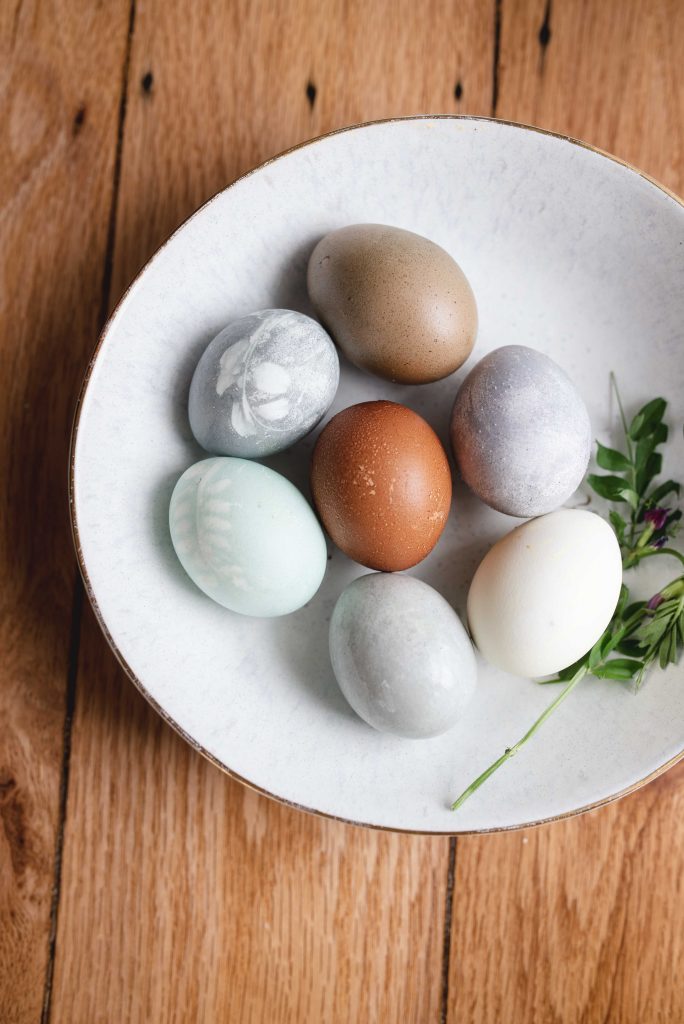 a bowl of vegetable dyed easter eggs with a sprig of greenery on a wooden counter