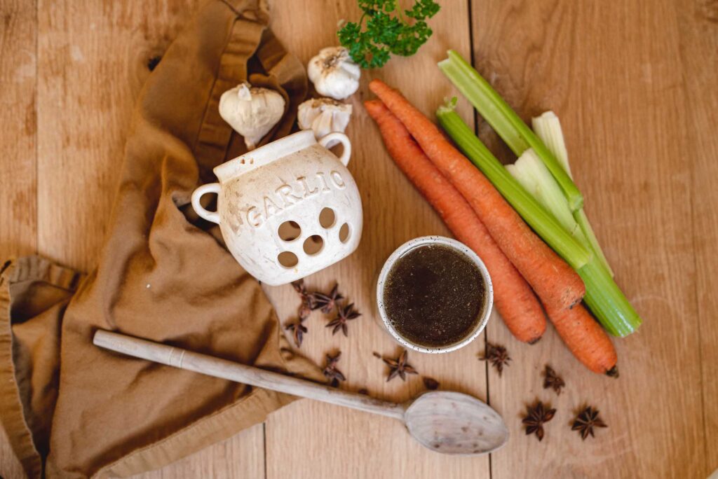 a wooden counter with an arrangement of bone broth materials: carrots, celery, garlic bulbs, parsley, and a stoneware cup of bone broth