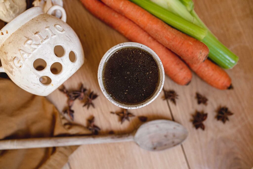 a cup of bone broth on a butcher block with carrots, celery, and star anise and a wooden spoon