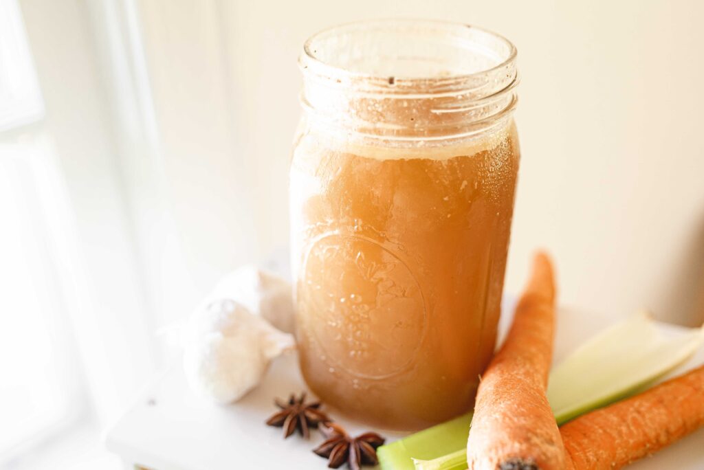 chicken bone broth in a mason jar next to a window with garlic bulbs, star anise, carrots, and celery scattered around